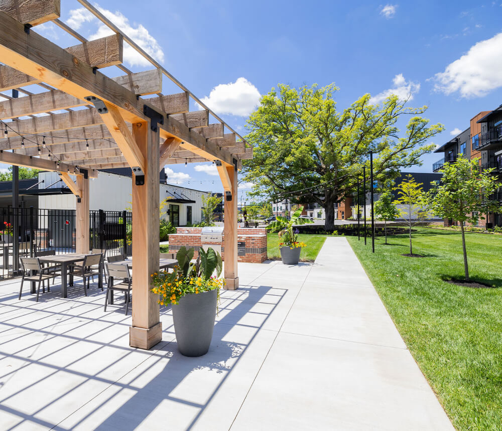 Covered cabana lounge over a concrete patio with dining tables and a large tree in a grassy courtyard.